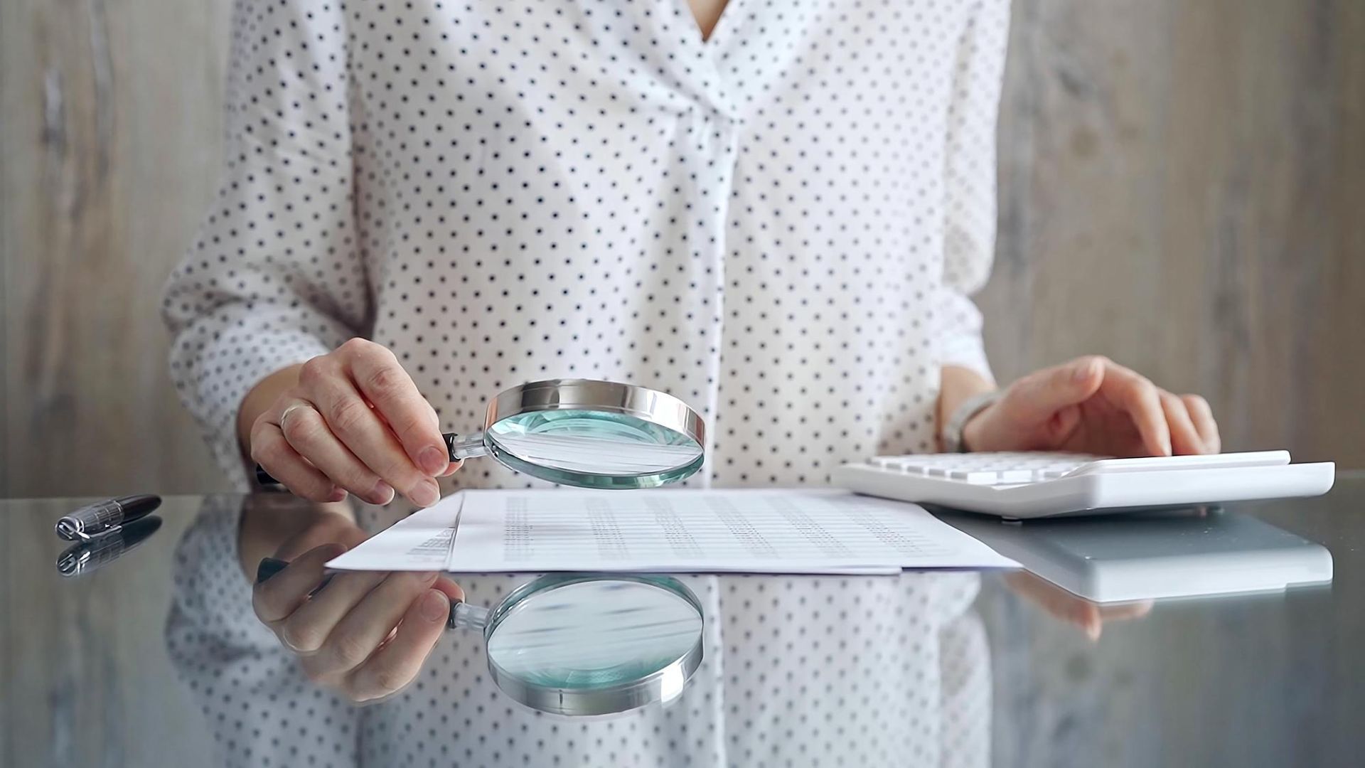 Woman examining document with a magnifying glass and using a calculator at a desk.