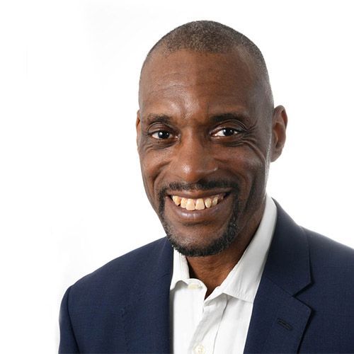 Man in navy blazer smiles, facing camera, against white background.