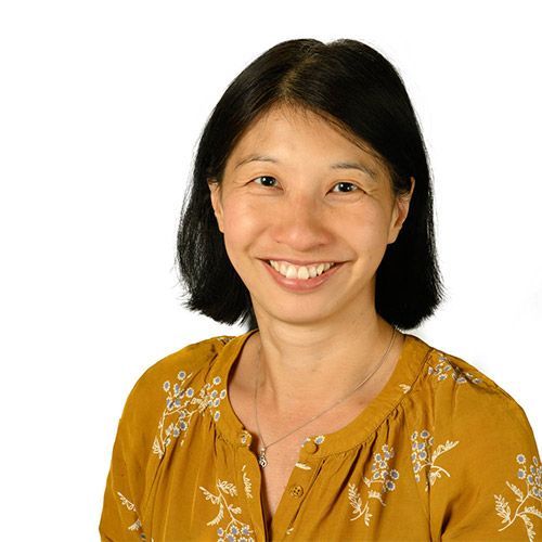 Woman with short black hair, smiling, wearing a mustard-colored blouse, against a white background.