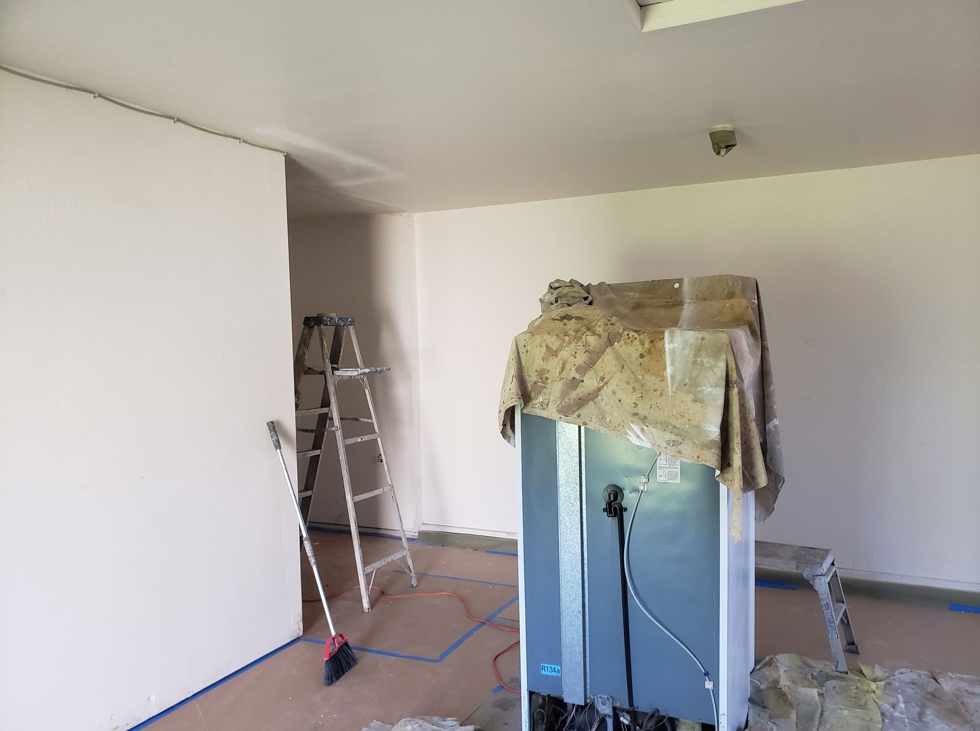 A refrigerator is sitting in a room with a ladder and a broom.