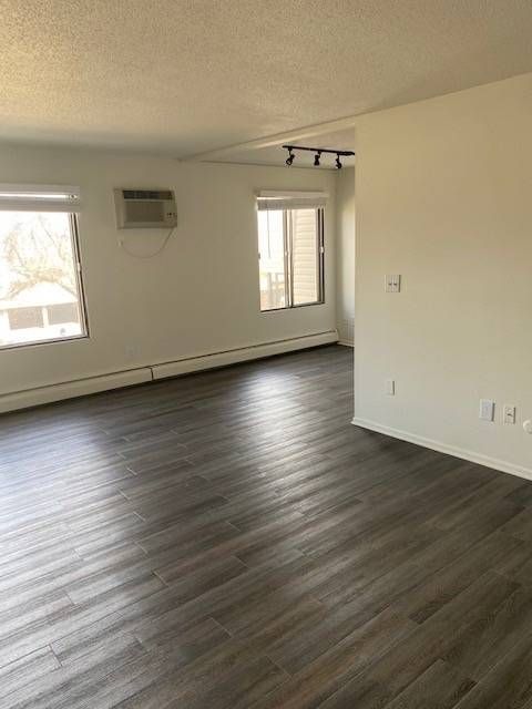 Empty living area with dark wood-look flooring, two windows, and a wall-mounted AC unit.