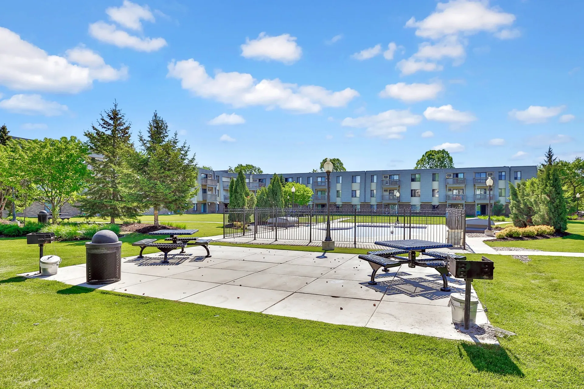 Park with picnic tables and grill in front of a multi-story building on a sunny day.