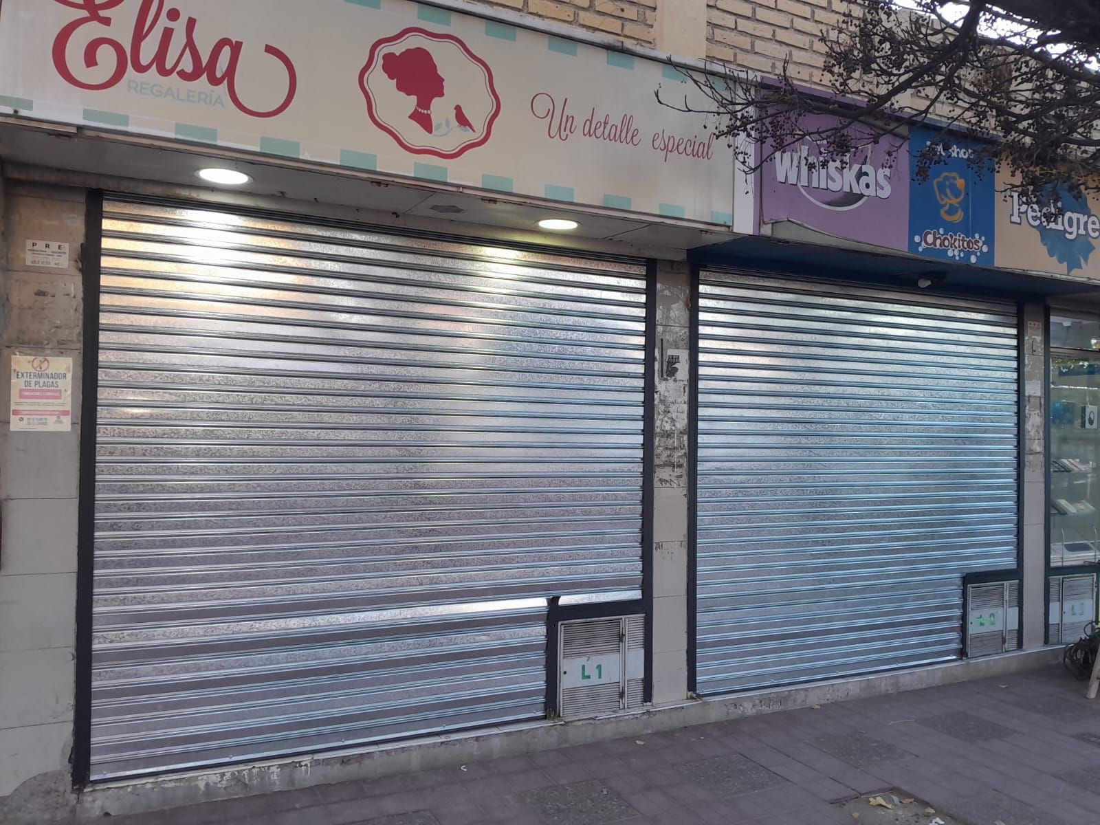 Tiendas cerradas con contraventanas metálicas plateadas y letreros descoloridos en la acera.