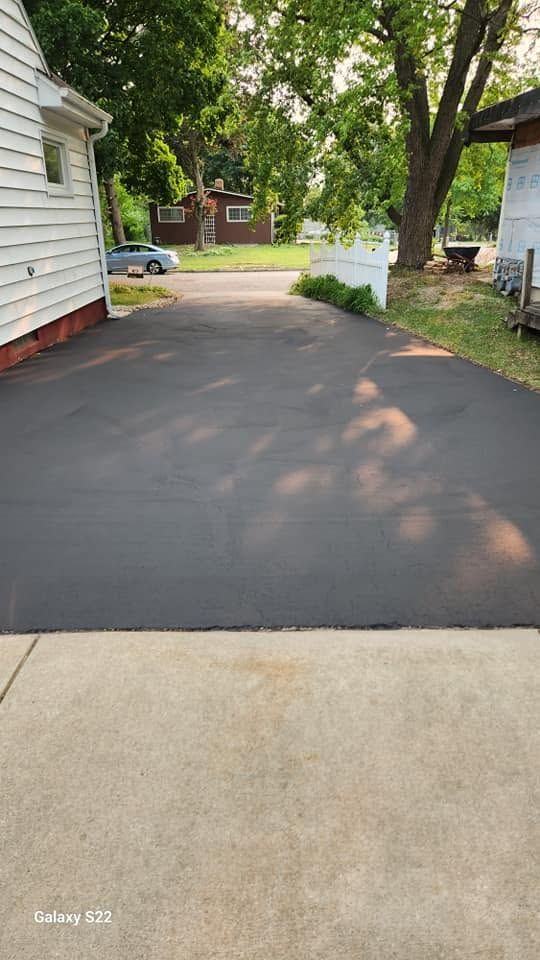 Freshly paved black driveway beside a white house, with trees and a quiet residential street in the background