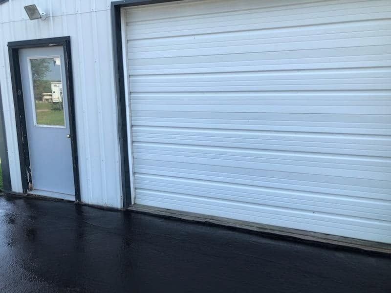 White garage door beside a gray entry door on a wet pavement outside an industrial building