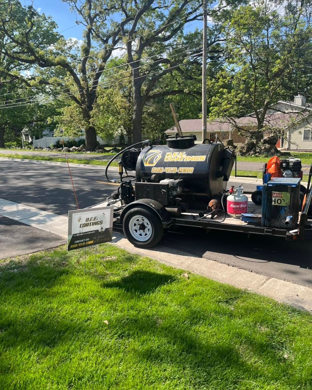 Lawn care trailer with black tank parked on a suburban street beside a green lawn and trees.