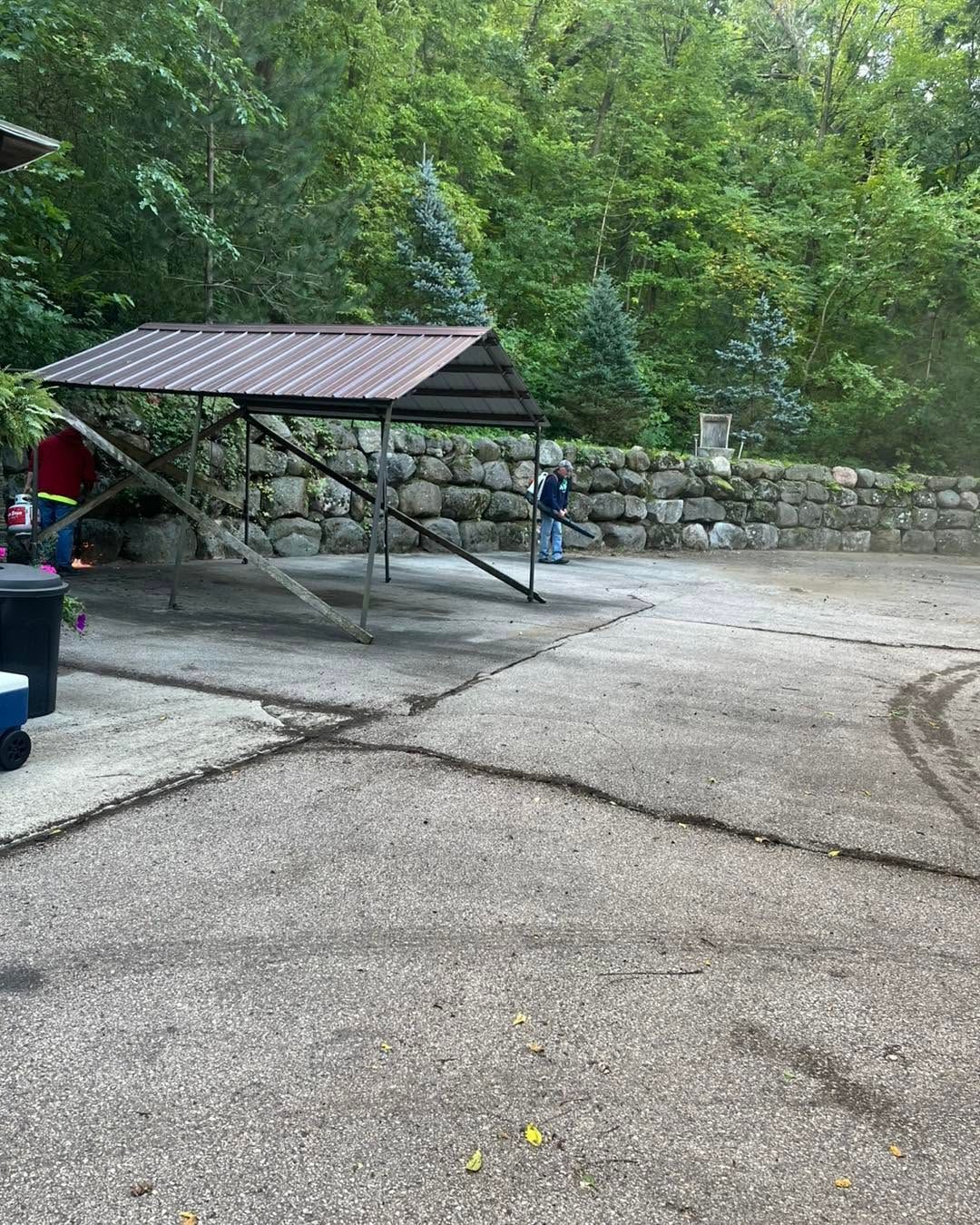 Outdoor paved area with a picnic shelter, stone wall, and a person walking near trees
