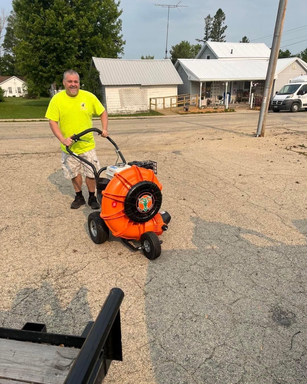 Man in neon shirt pushing an orange leaf blower on a gravel driveway near houses