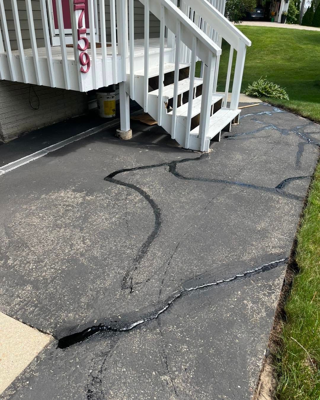 Cracked asphalt driveway beside white stairs leading to a porch and grassy yard