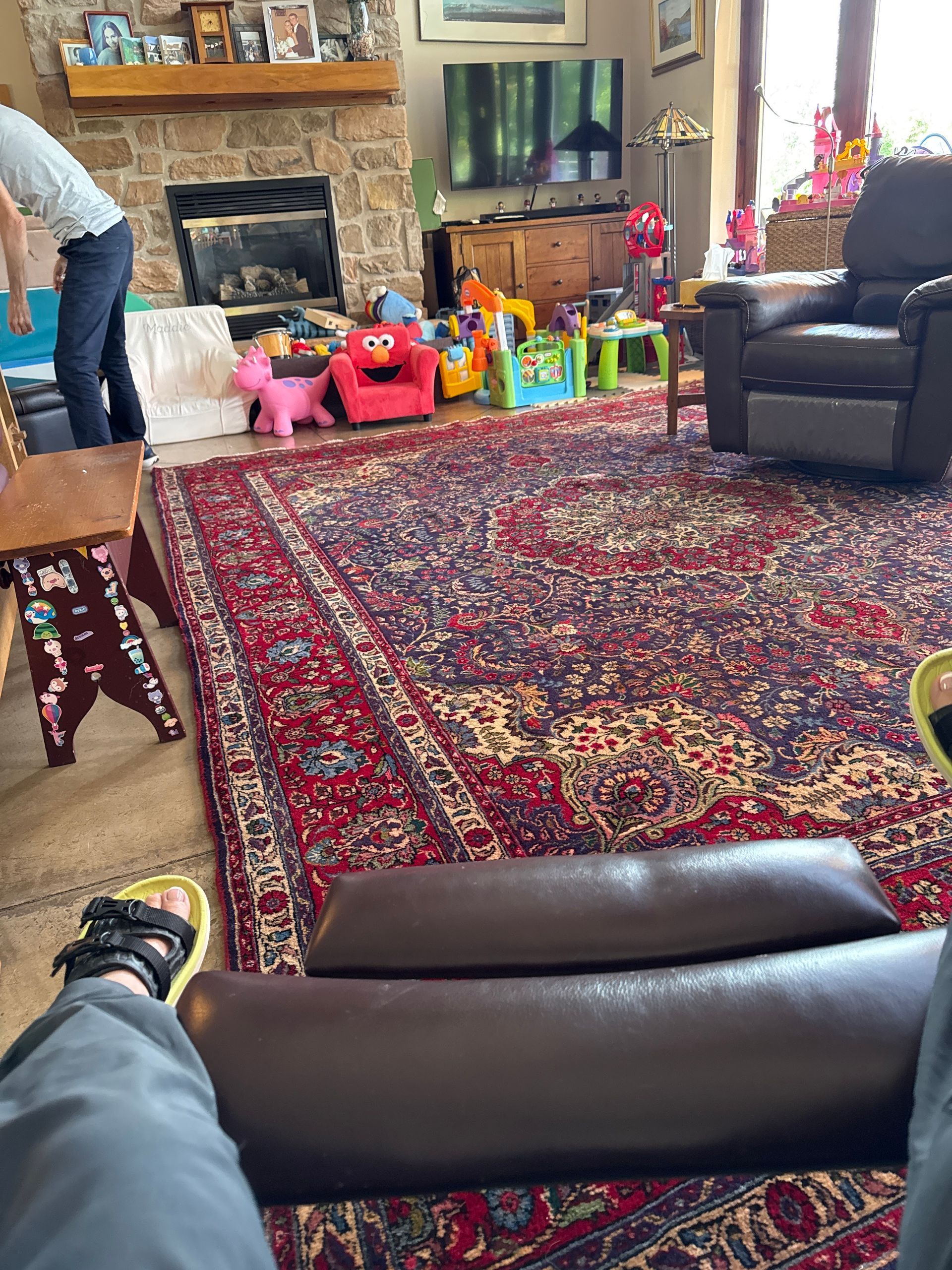 Person reclined in a brown leather chair, looking at a living room with toys and fireplace, large patterned rug.