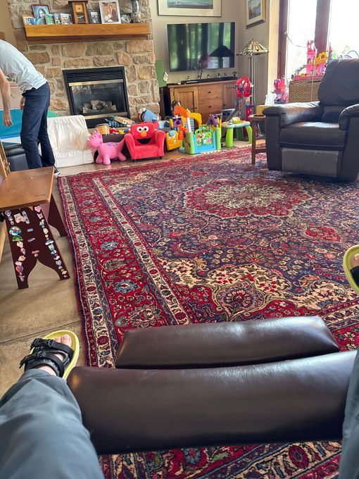 Person in recliner views living room with fireplace, toys, and oriental rug.