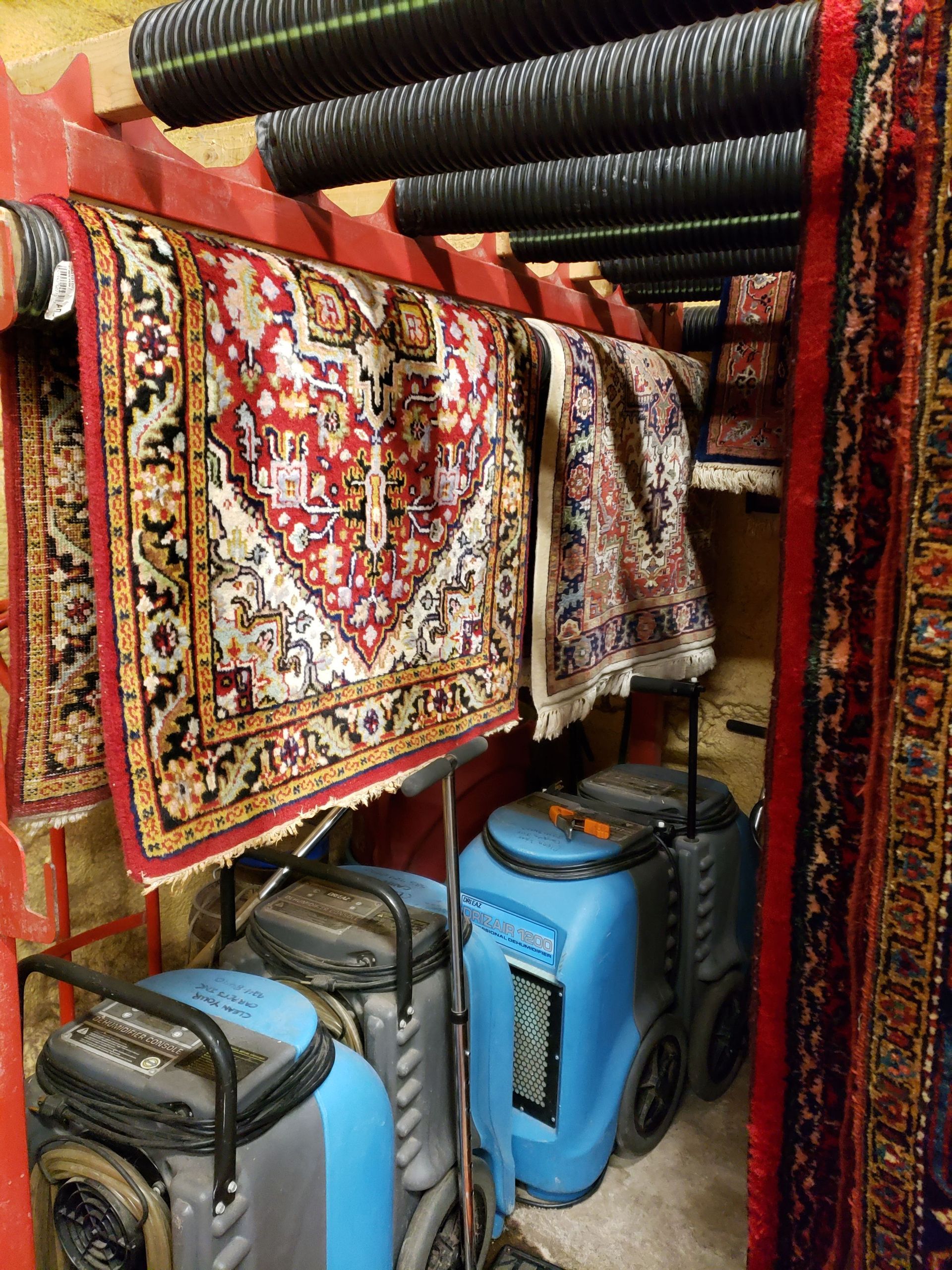 Rugs hanging to dry over dehumidifiers in a storage area. Red and beige patterns, blue machines.