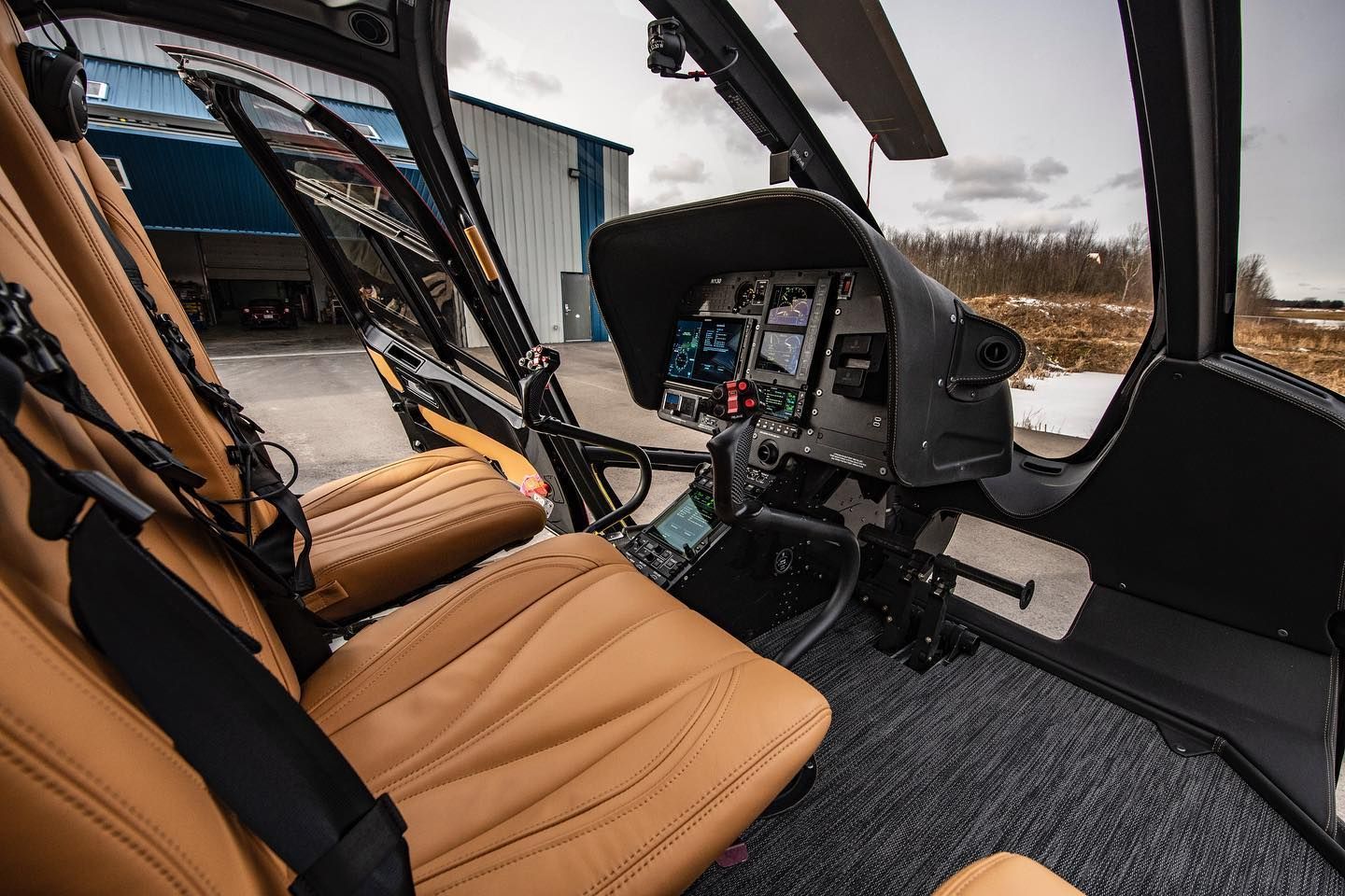 Interior view of a helicopter cockpit with tan leather seats and a digital instrument panel. The door is open.