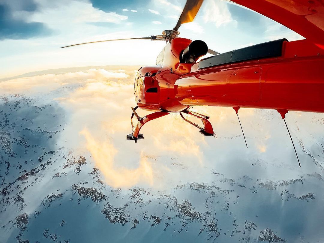 Red biplane soaring above snow-covered mountains, bright sky.