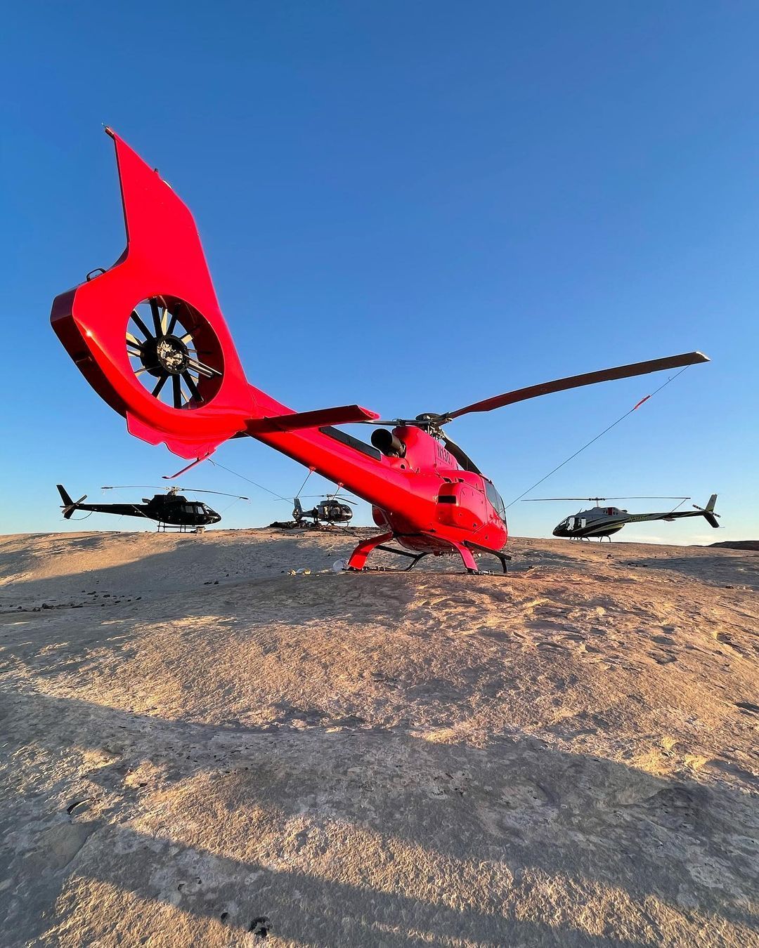 Red helicopter on a rocky surface with three black helicopters in the background under a clear blue sky.