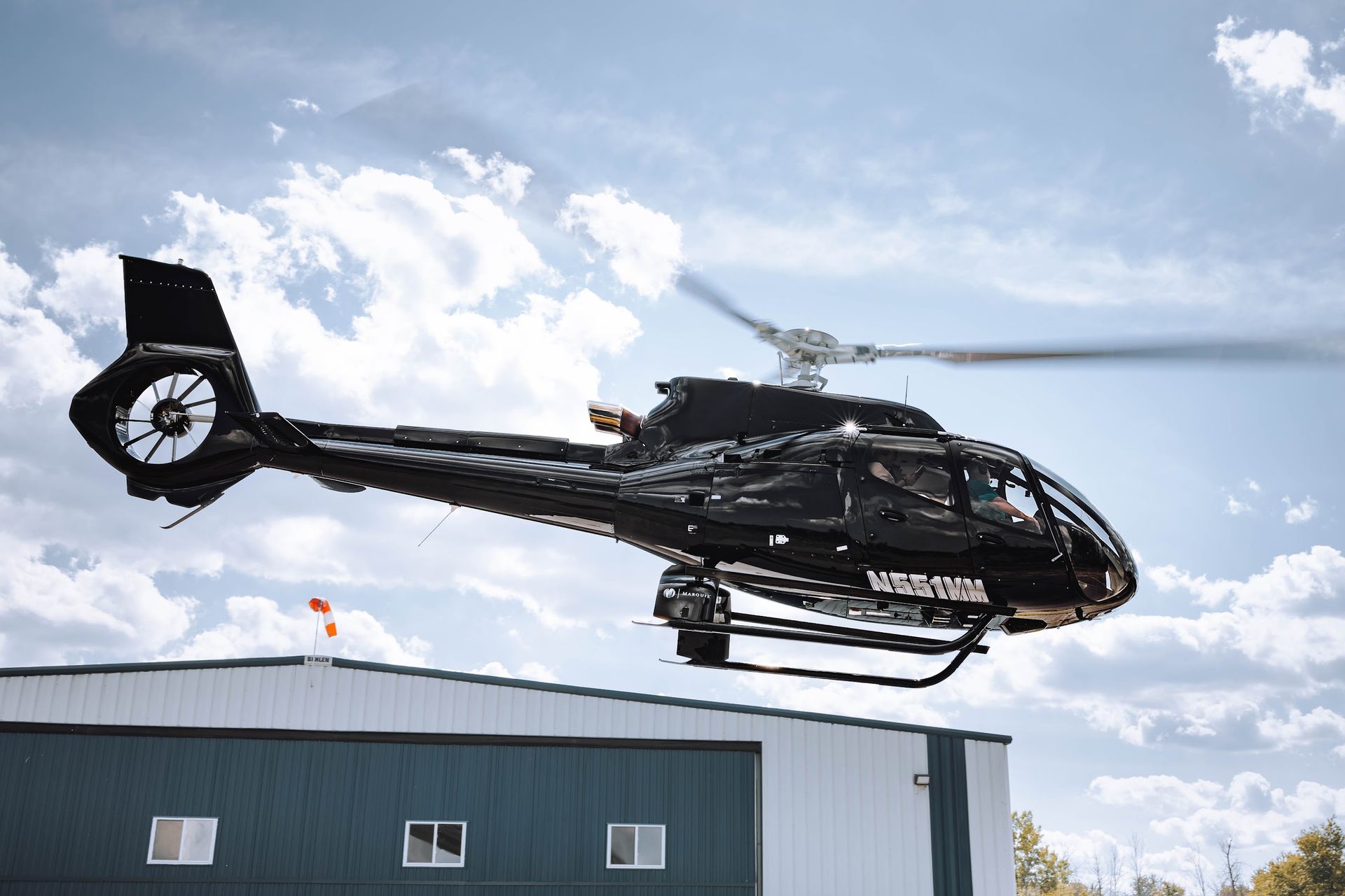 Black helicopter taking off near a building with a blue and white exterior, against a cloudy sky.