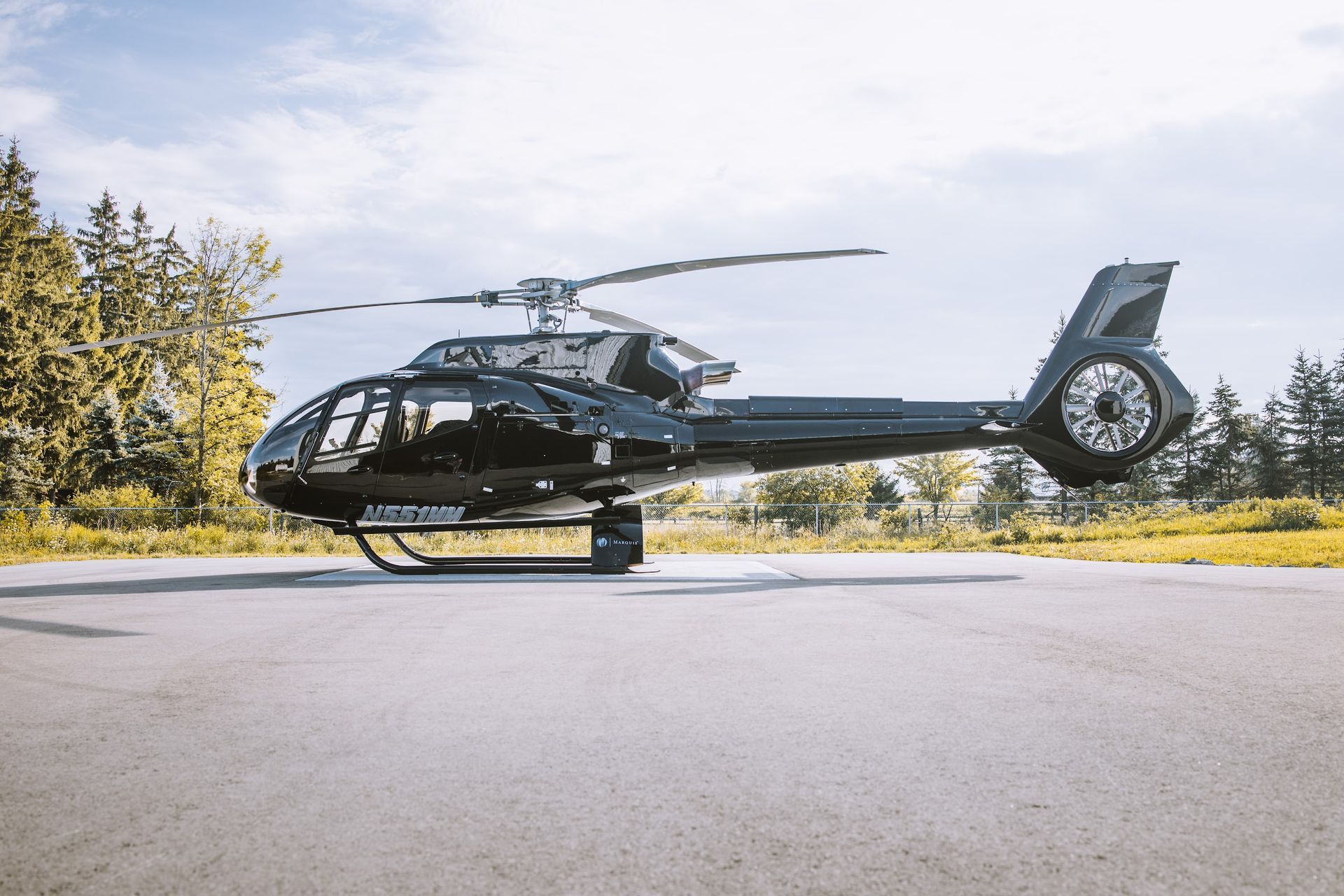 Black helicopter on a concrete pad with trees in the background under a blue sky.
