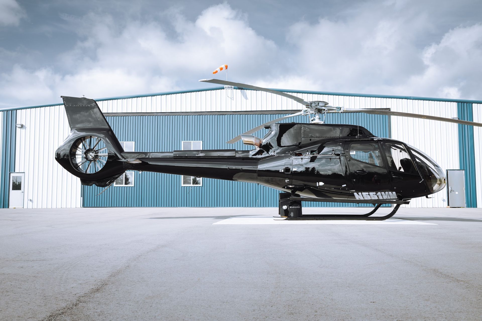 Black helicopter on a tarmac in front of a blue and white building, cloudy sky above.