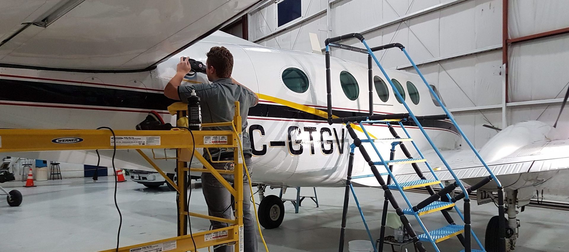 A person working on an airplane in a hangar. They stand on a yellow scaffold near the aircraft's side.