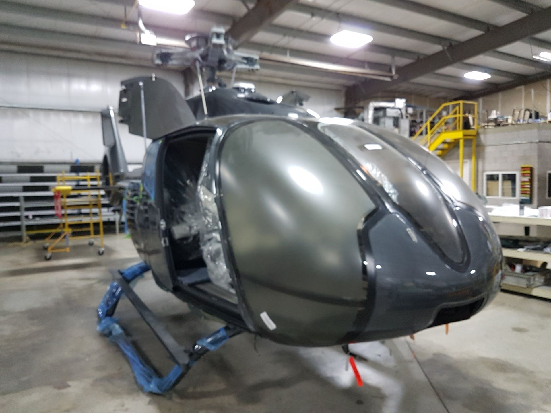 Dark gray helicopter in a hangar, door open. Blue protective wrapping on landing gear.