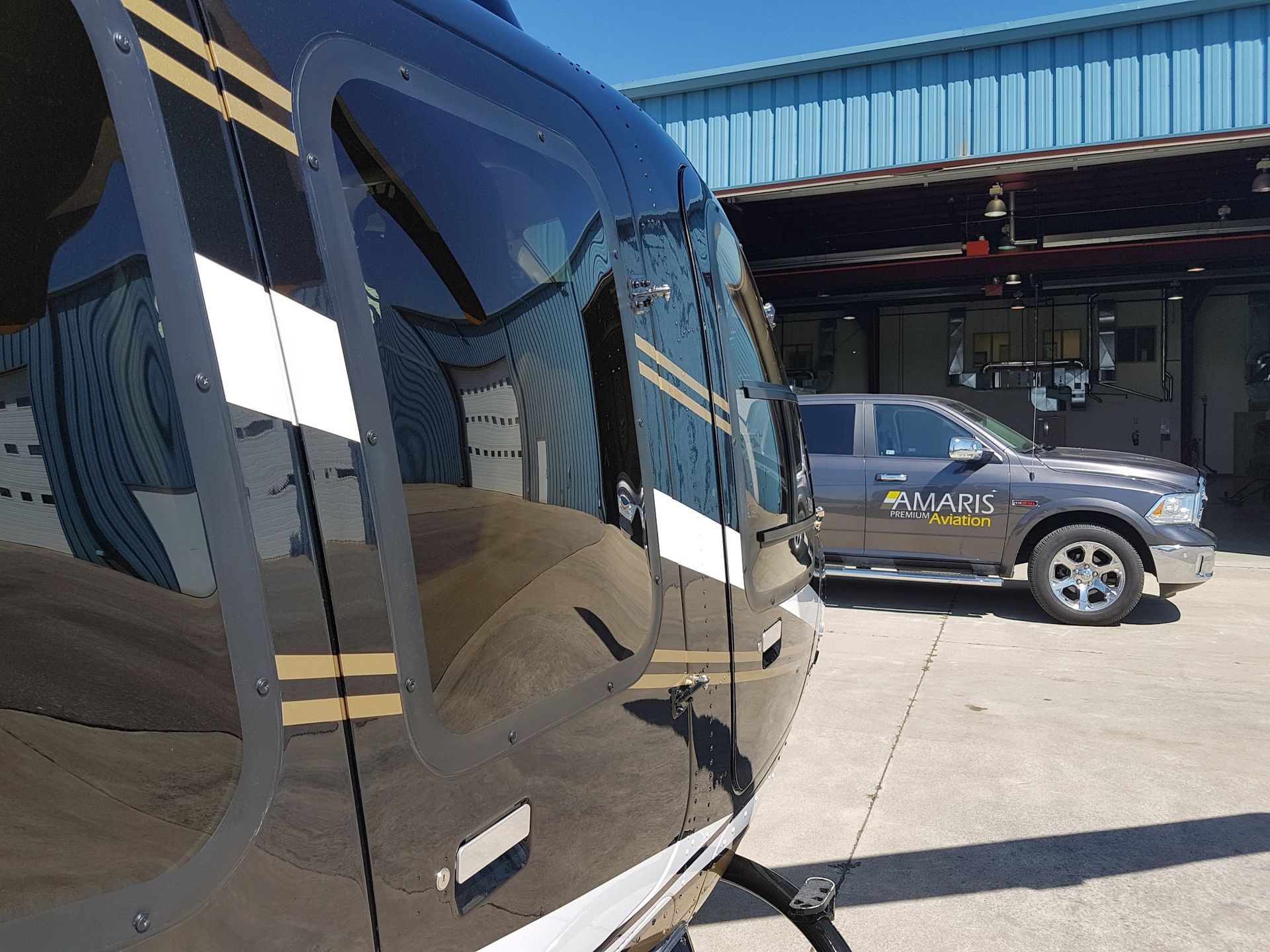 Black helicopter reflecting a building and a gray pickup truck parked outside a hangar on a sunny day.