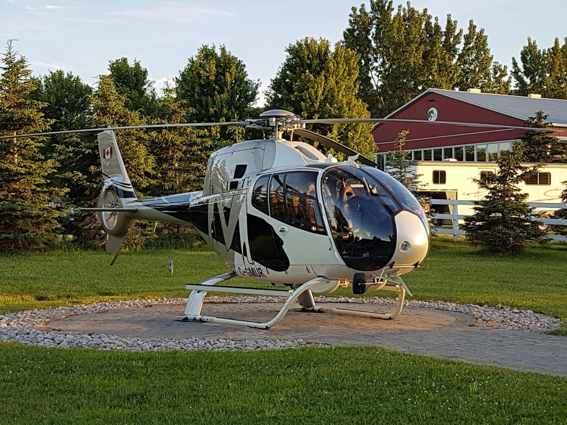 Helicopter on a circular pad of stones in a grassy area with trees and a red barn in the background.
