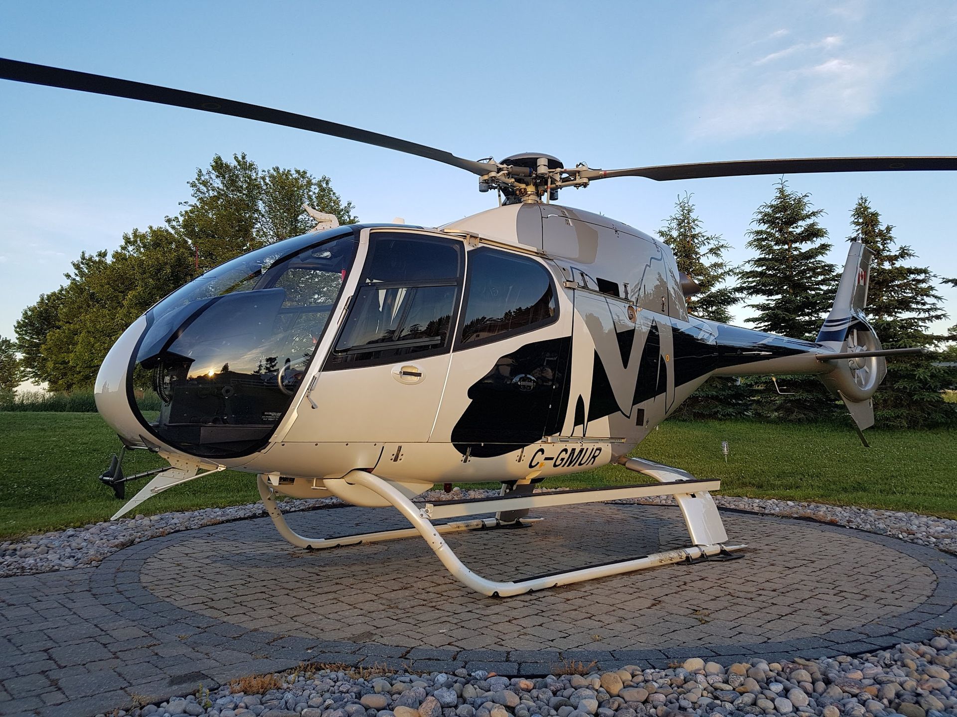 Helicopter on a circular pad, white and black camouflage paint, setting sun in the background.