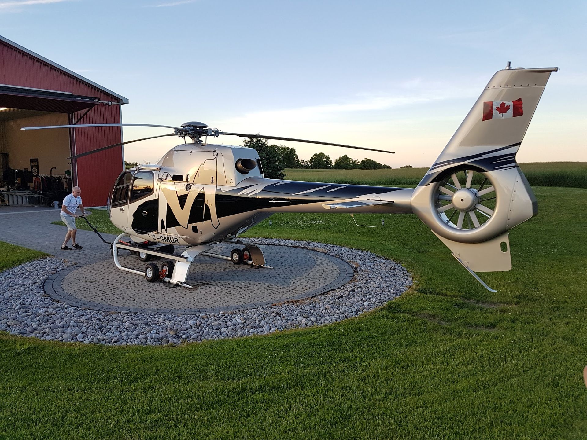 Helicopter on a stone pad, tail with Canadian flag. Man stands nearby. Building and grass in background.