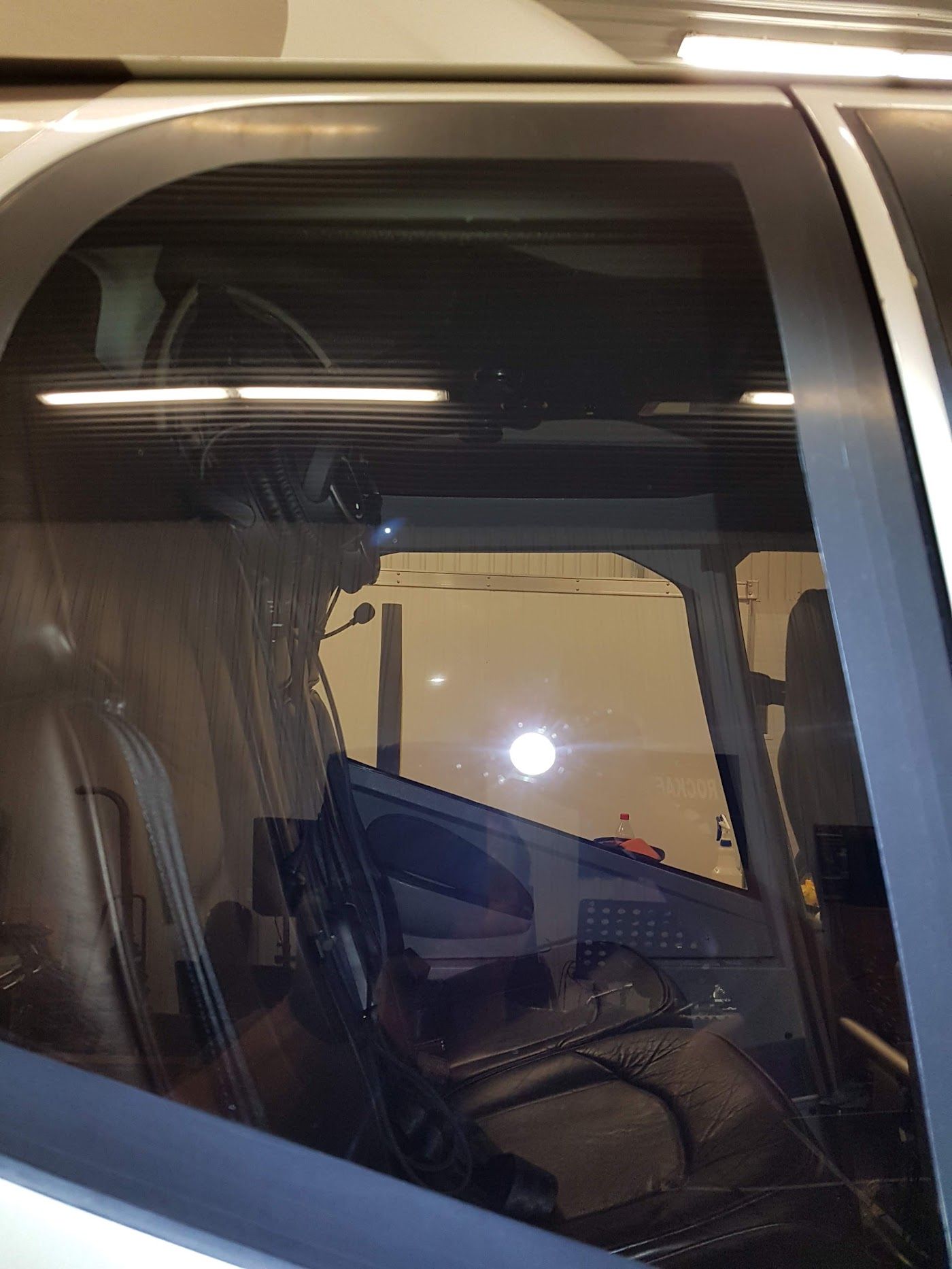 Interior view of a cockpit through a window, showing seats and a glimpse of a hangar with a bright light.