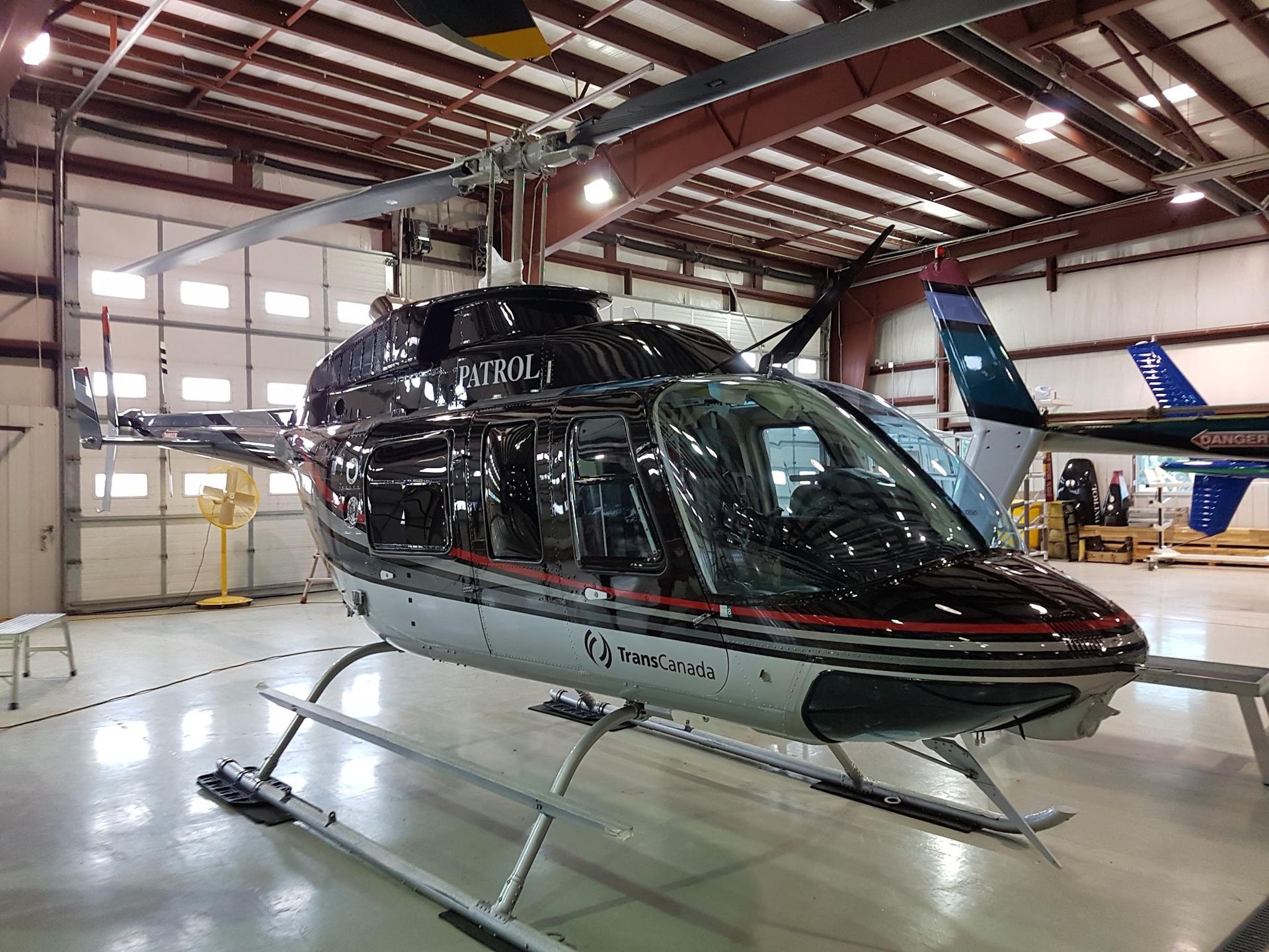 Black and silver helicopter inside a hangar with a partially open door, other aircraft visible in background.