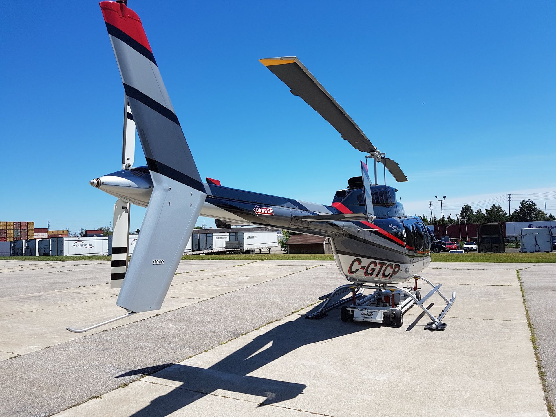 Helicopter on a tarmac with rotor blades extended. Tail fin is gray, black, and red; blue sky.