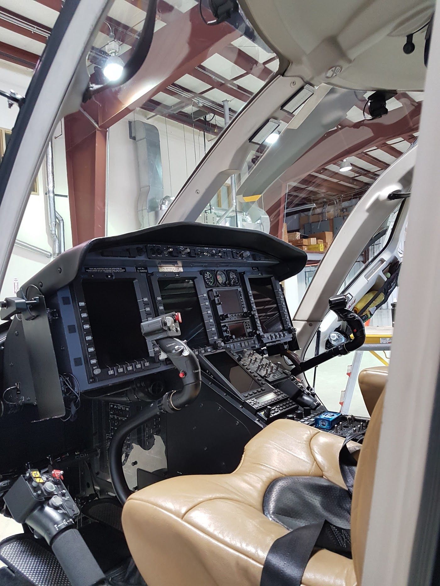 Helicopter cockpit interior: dark console with screens, tan seat, and overhead structure, likely in a hangar.