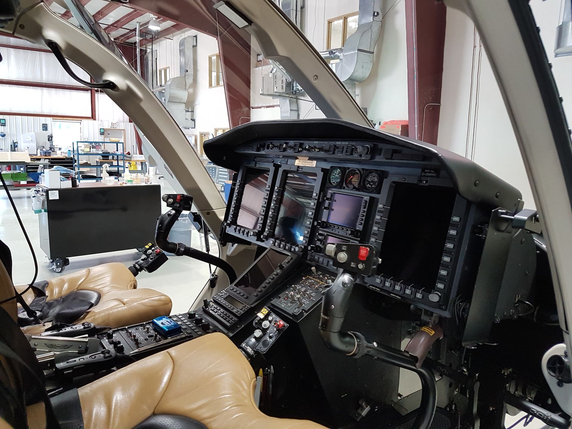 Helicopter cockpit interior view: displays, controls, tan seats. Industrial setting.