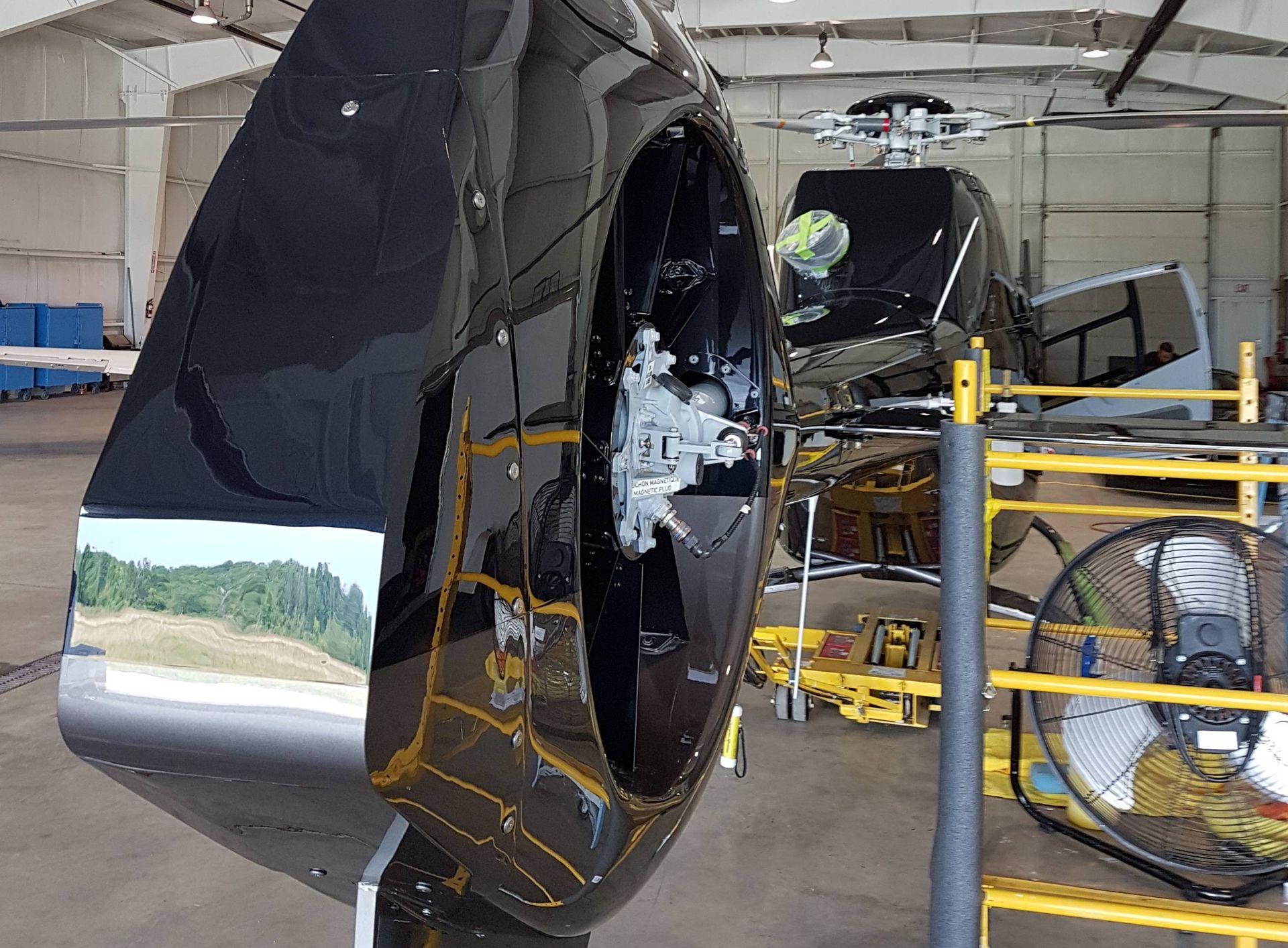 Black helicopter tail rotor inside a hangar; reflecting green trees and a blue sky.