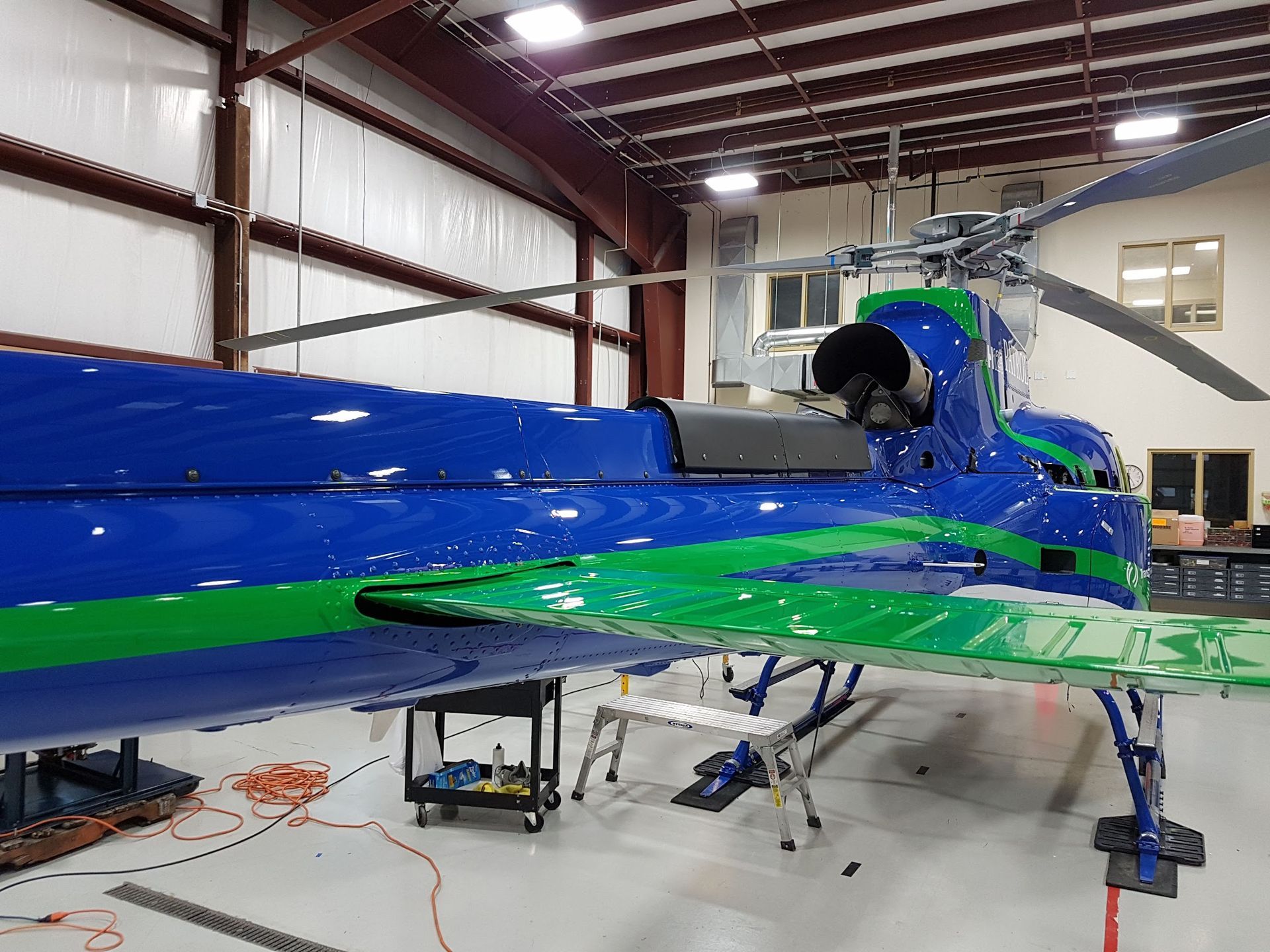 Blue and green helicopter in a hangar with the tail visible, rotors extended.