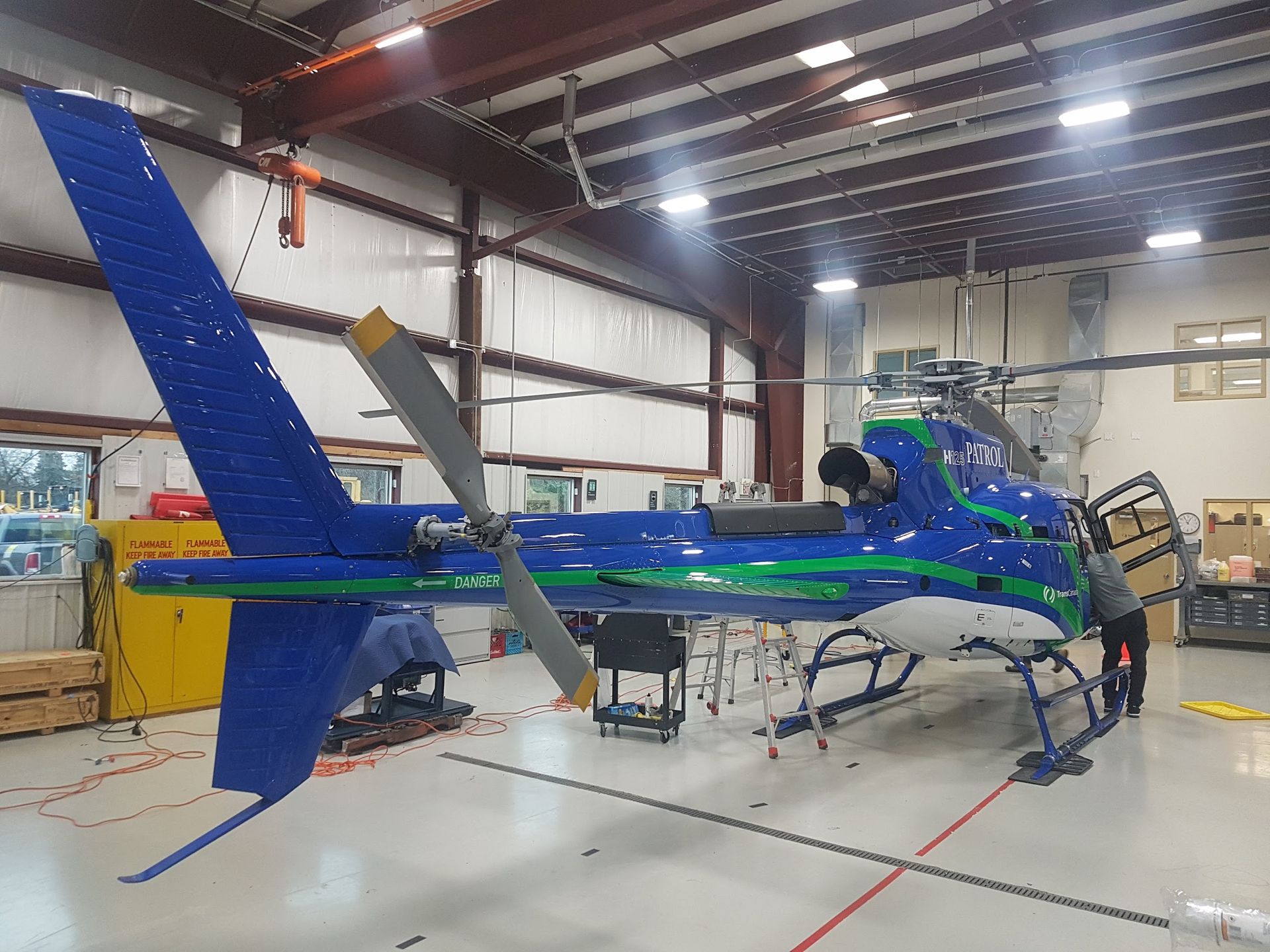 Blue and green helicopter in a hangar, being worked on by two people.