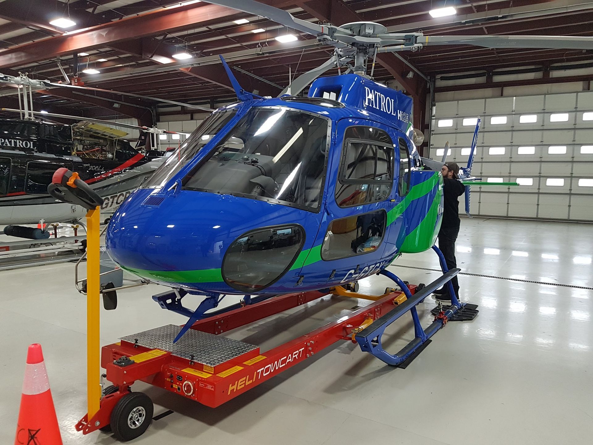 Blue and green helicopter on a red maintenance cart inside a hangar. A person is nearby.
