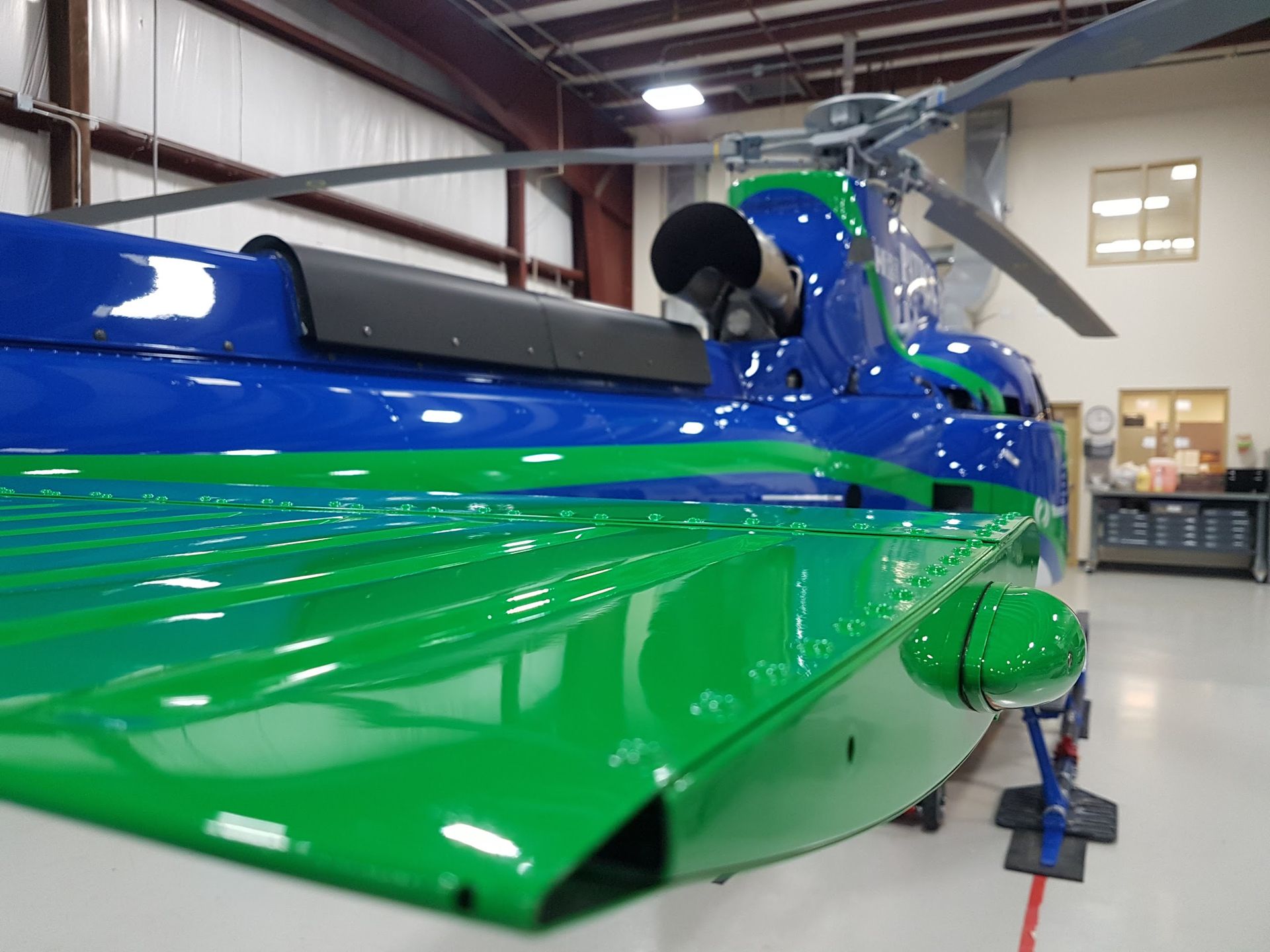 Green and blue helicopter in a hangar, angled view of side.