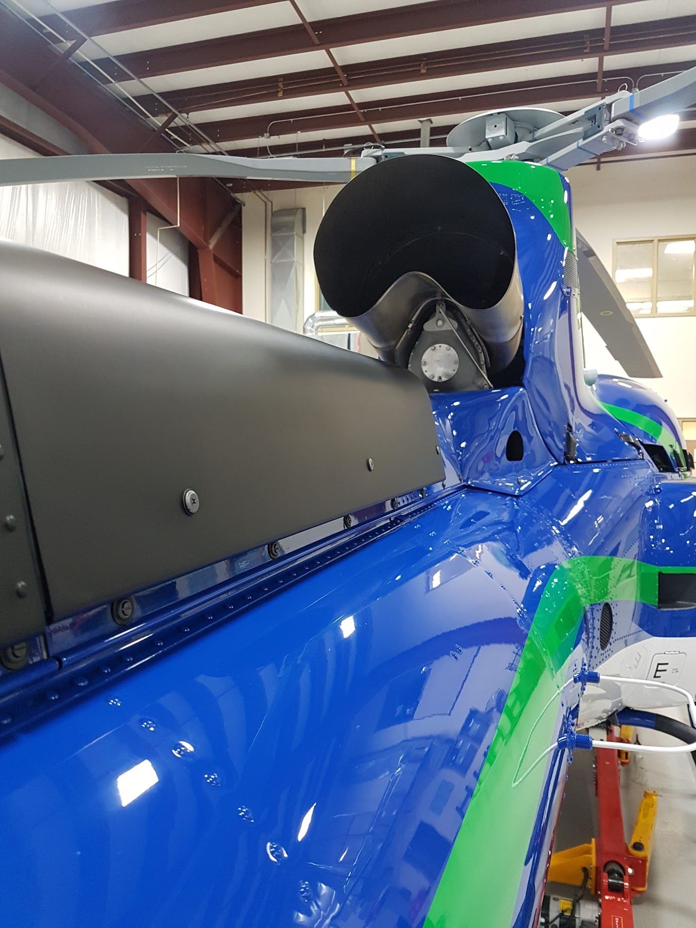 Blue and green helicopter body with a black exhaust pipe in a hangar.