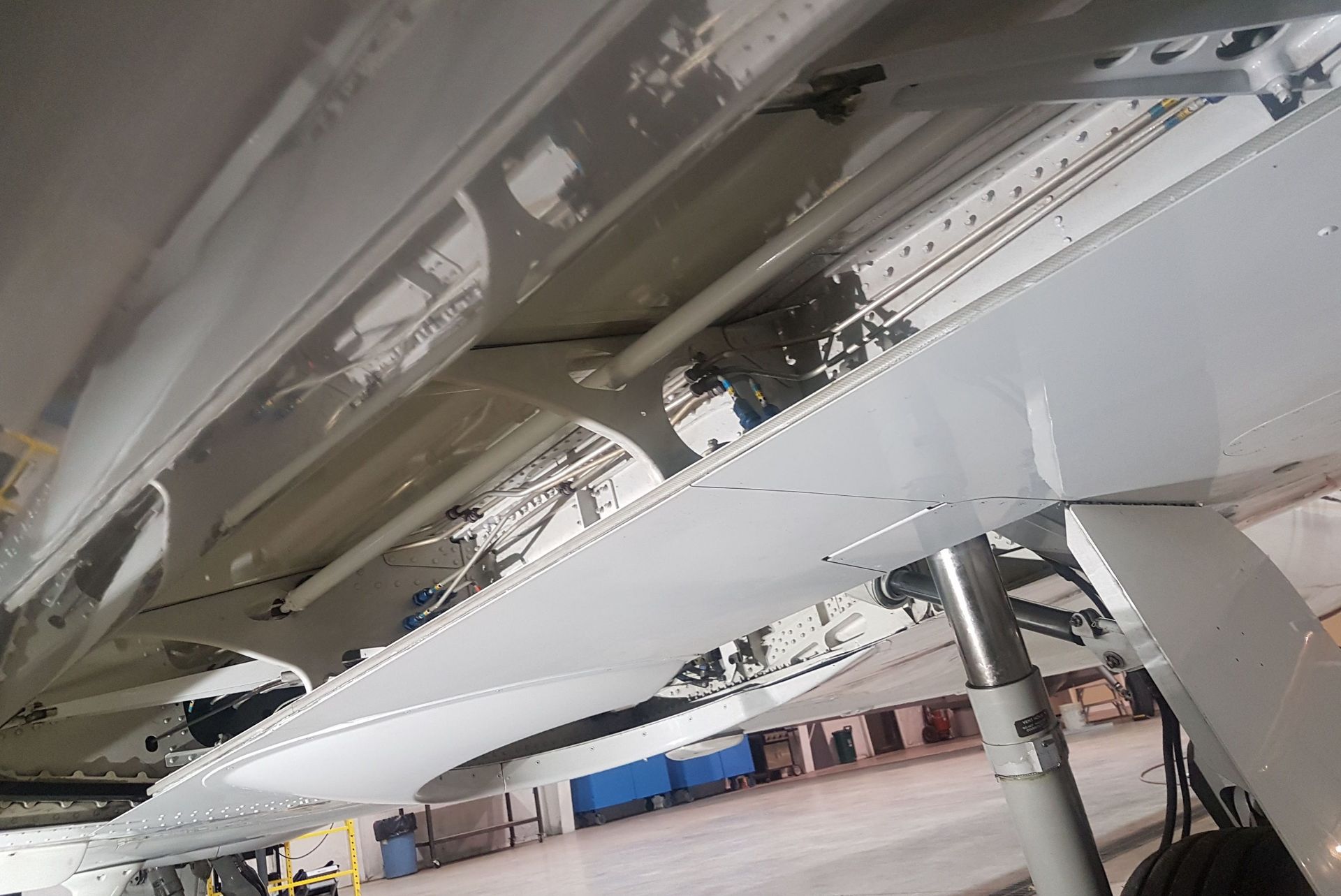 Underside of an airplane wing. White metal framework with support beams, visible wiring, and a landing gear strut.