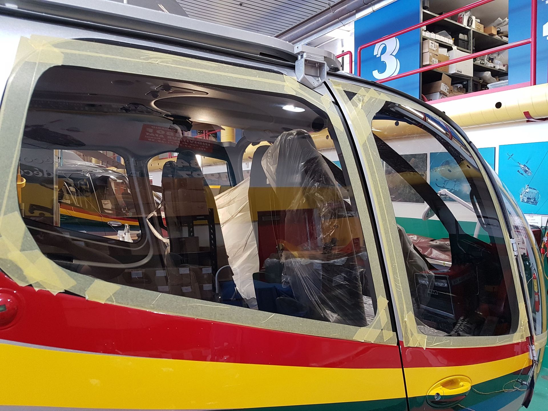 Helicopter cockpit interior partially masked for painting; red, yellow, and green exterior paint visible.