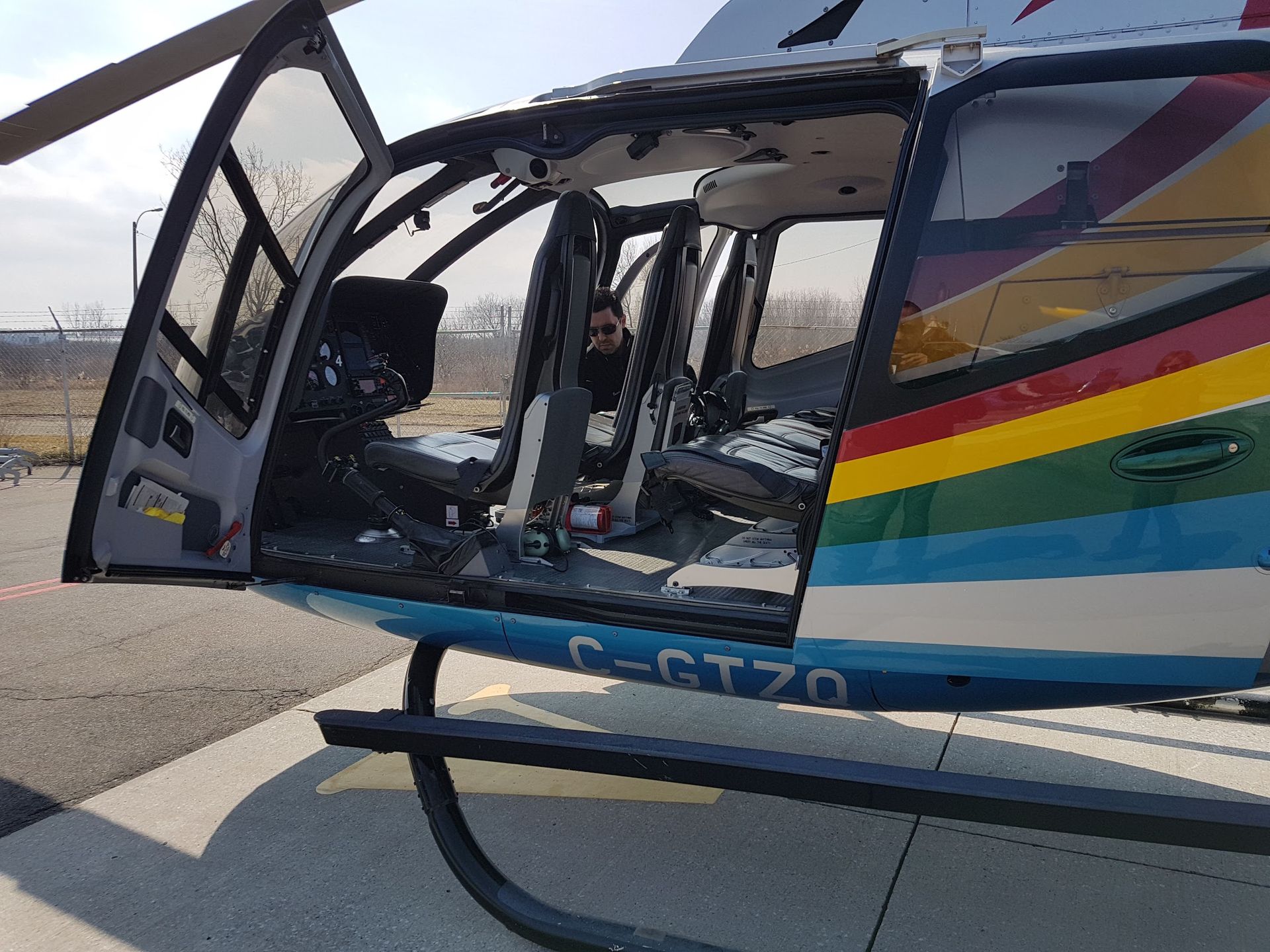 Helicopter with open doors; multi-colored stripe design, seated pilot visible.