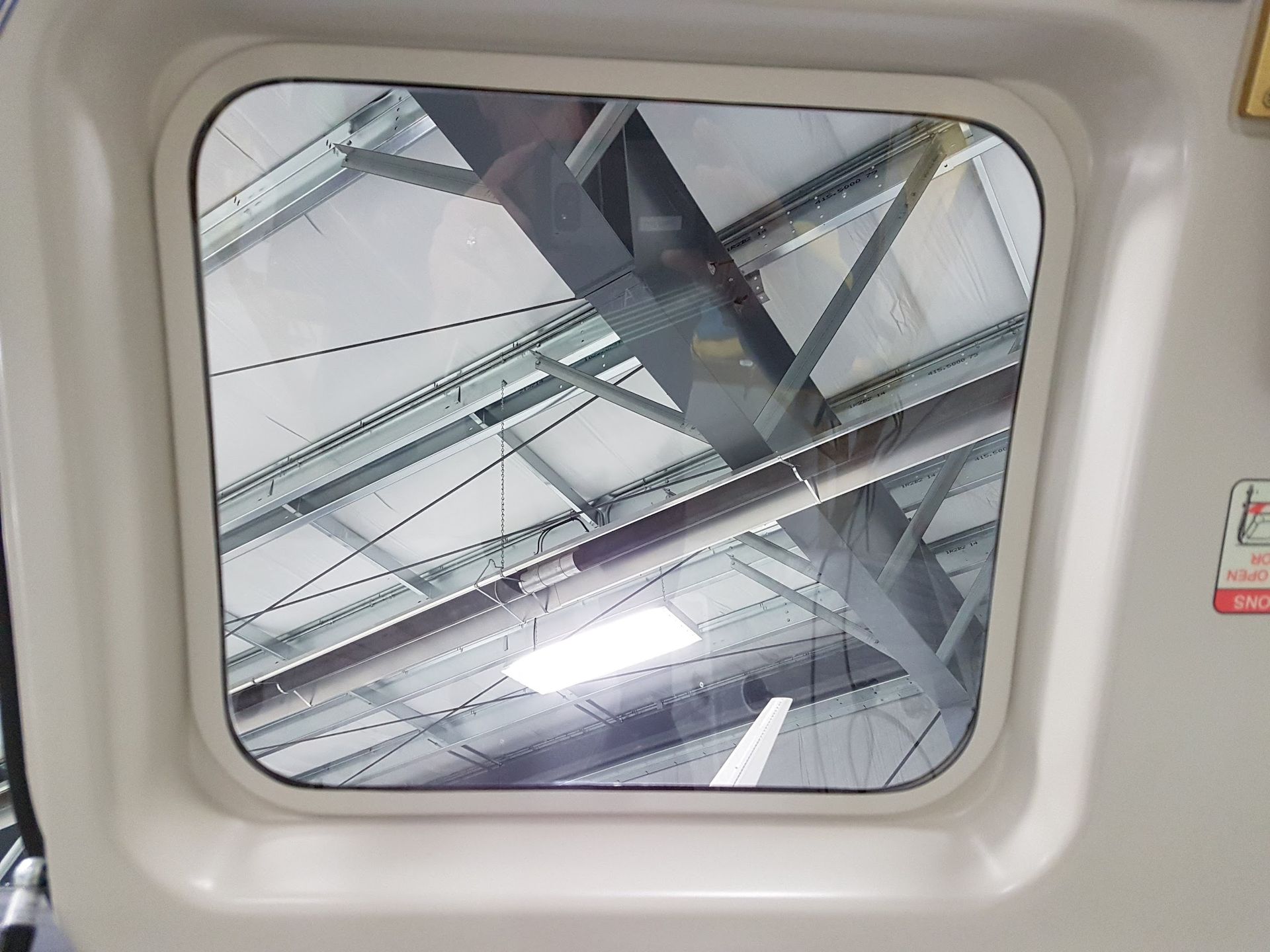 Window showing a warehouse ceiling with beams, pipes, and lights.