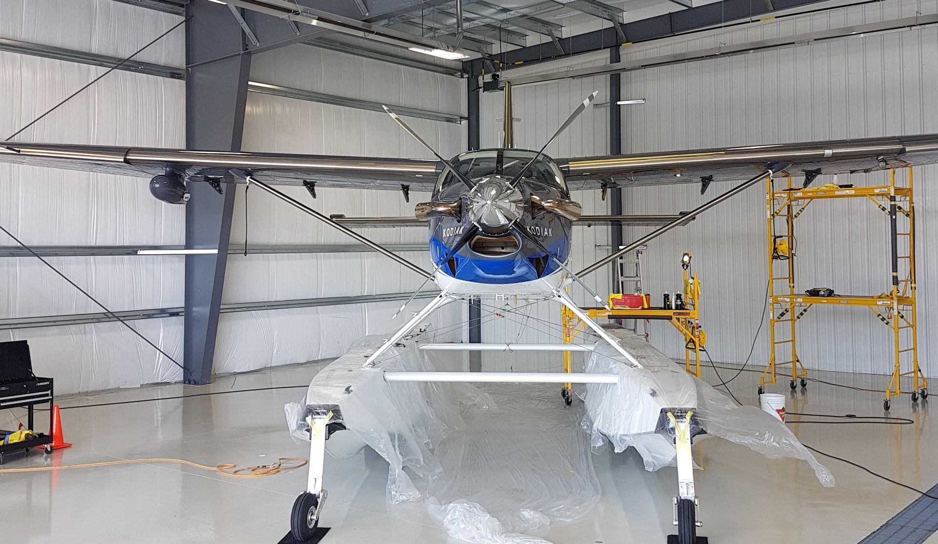 Airplane in a hangar, supported by scaffolding. The plane has a blue nose and gray wings.