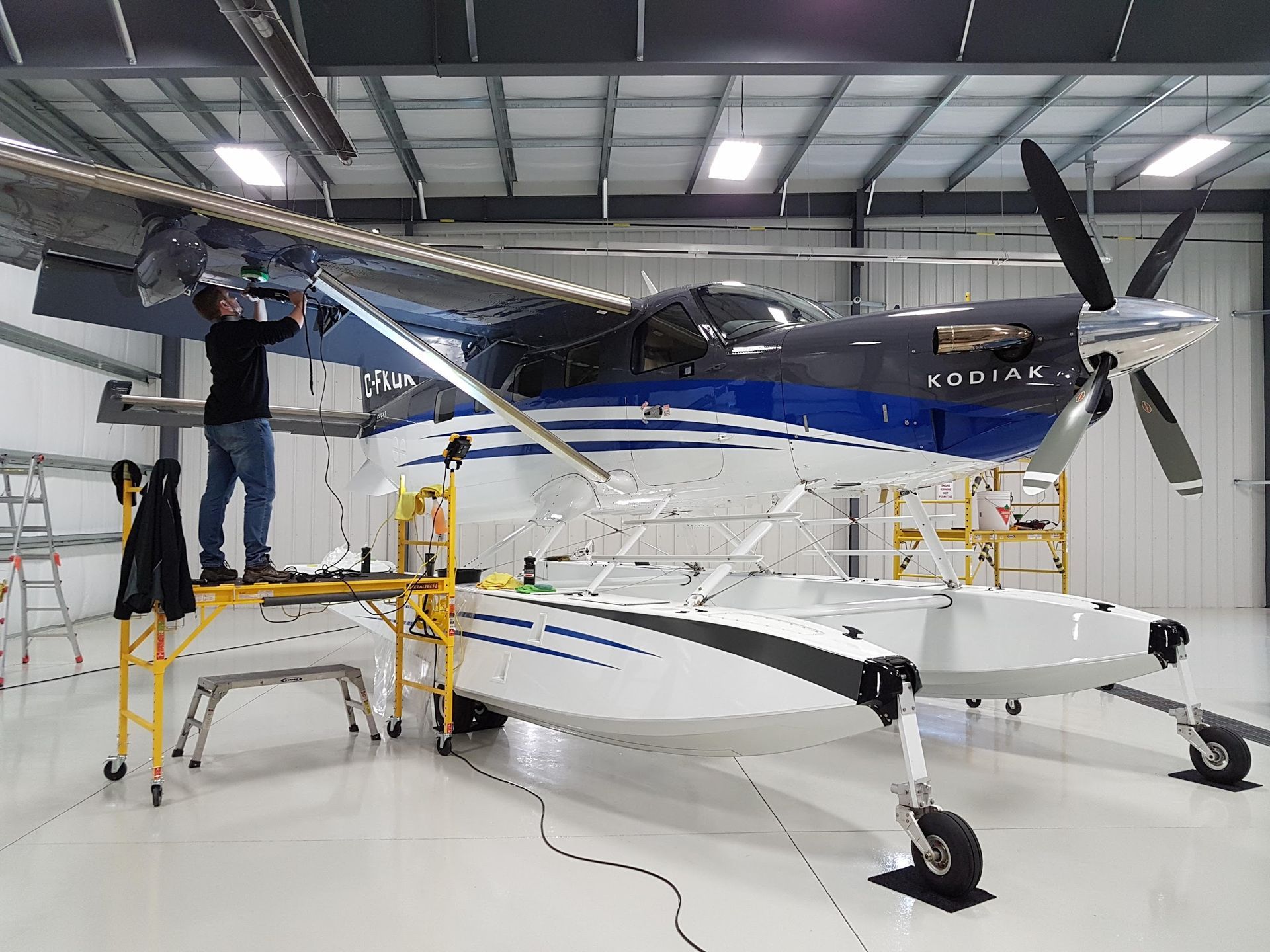 Man working on a seaplane in a hangar; the plane is white and blue, with floats.