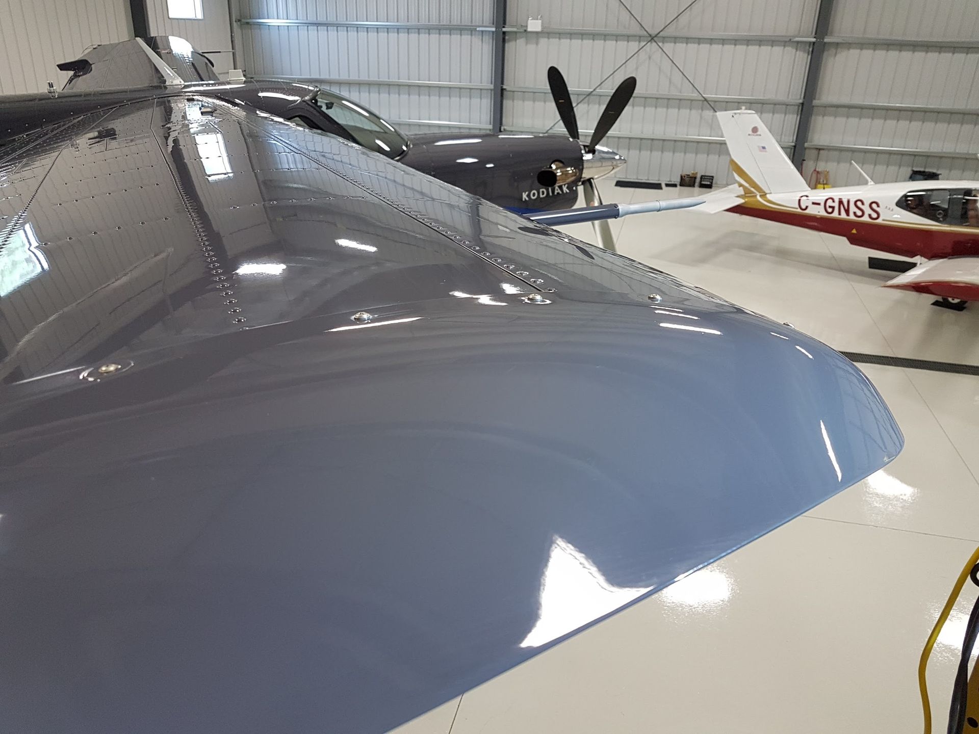 Wing of a gray aircraft inside a hangar, with other planes in the background.