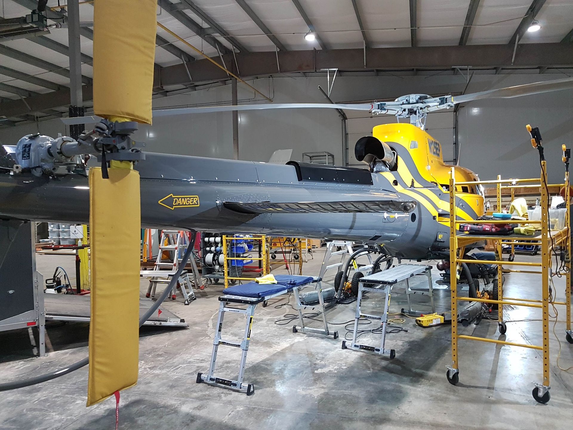 Helicopter inside a hangar, undergoing maintenance. Yellow and black accents, scaffolding, tools.