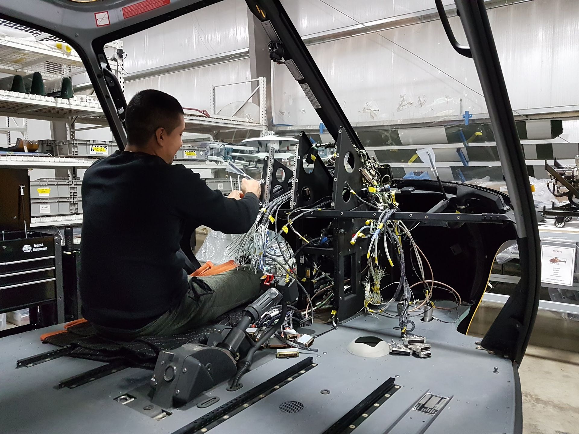 A person working on the interior of a helicopter, connecting wires. Setting is an industrial workshop.