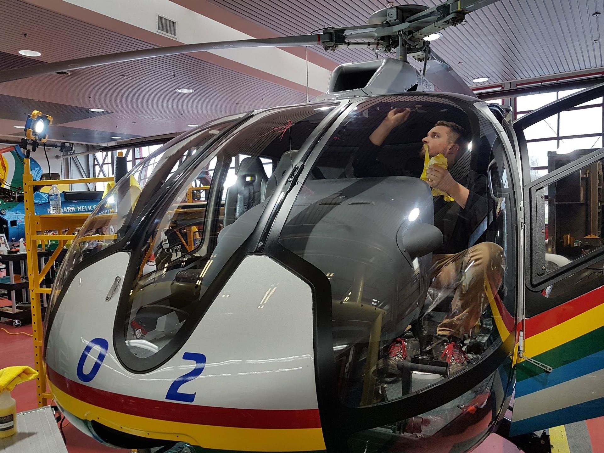 Man cleaning the inside of a gray and white helicopter. Interior shot of the cockpit with a rainbow stripe on the side.