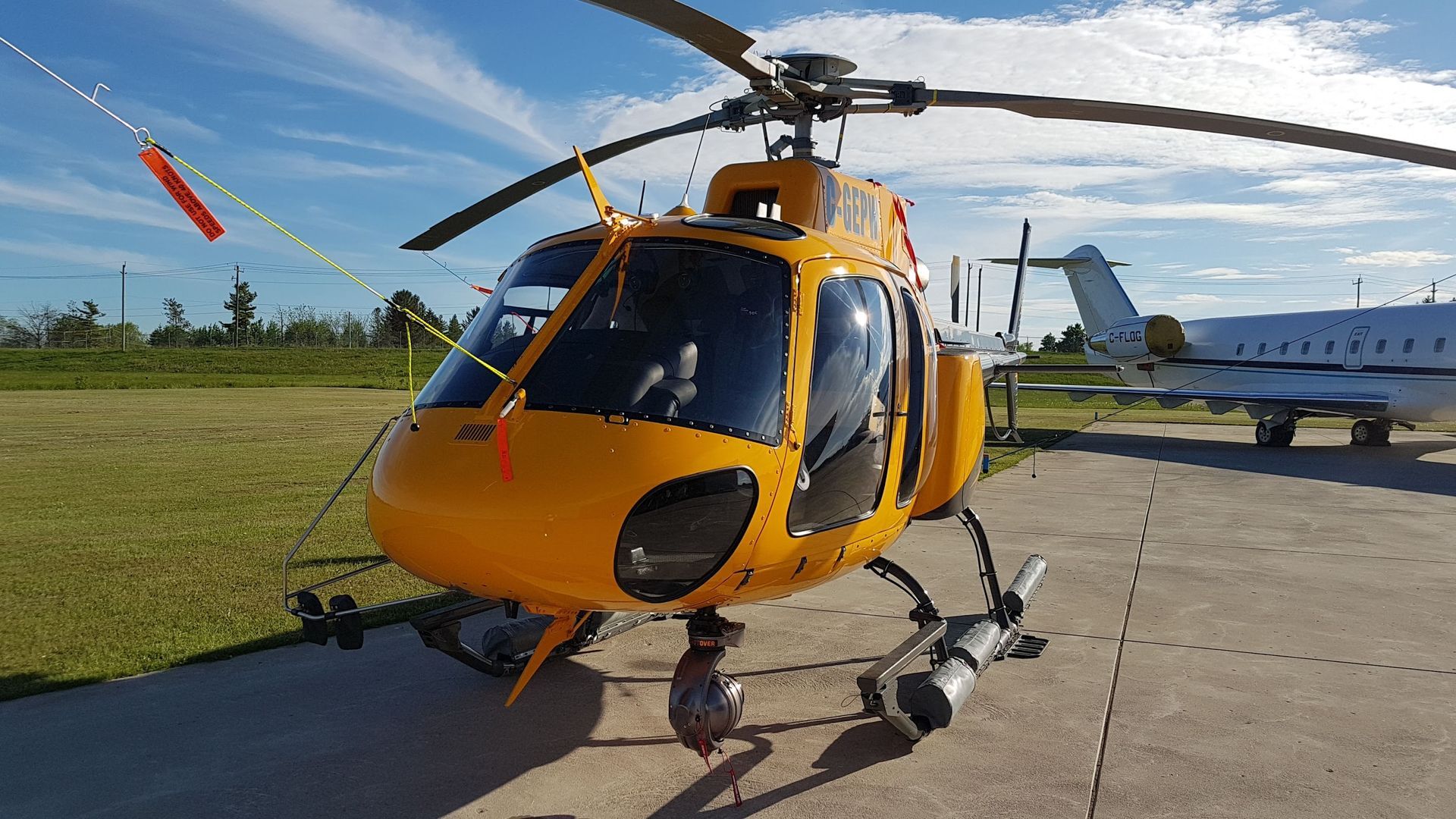 Yellow helicopter parked on a tarmac, sky background, with a plane visible in the distance.